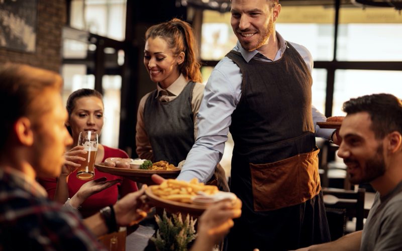 Happy waiters bringing food at the table and serving group of friends in a restaurant.